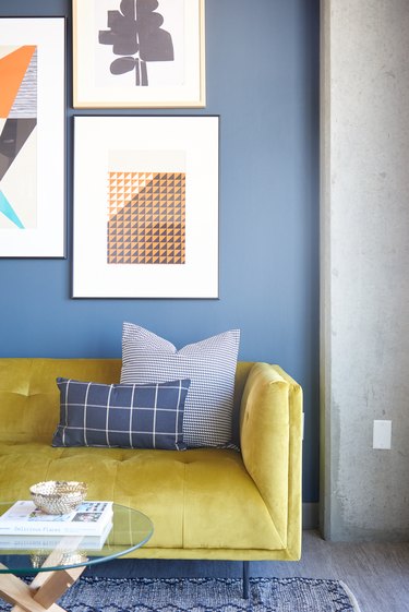Eclectic living room with yellow couch, blue wall, art, pillows, round glass coffee table, blue rug.